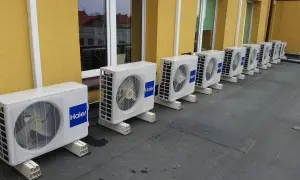 Rząd białych jednostek klimatyzacyjnych Haier na dachu, na tle żółtej ściany budynku.