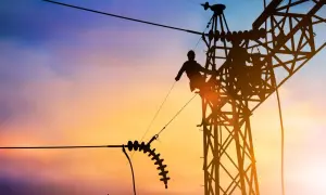 Sylwetka pracownika na tle zachodzącego słońca, naprawiającego linię energetyczną. Widać izolatory i konstrukcję wieży, a także potencjalny łuk elektryczny.