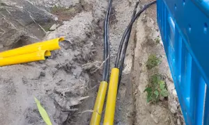 Wykop pod instalację światłowodową. Widoczne żółte osłony kabli i czarne przewody w wykopie. Obok niebieskie ogrodzenie budowlane.