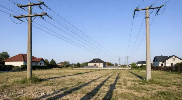 Dwa słupy średniego napięcia przecinają niebo nad pustą działką. W tle widać domy i drzewa.