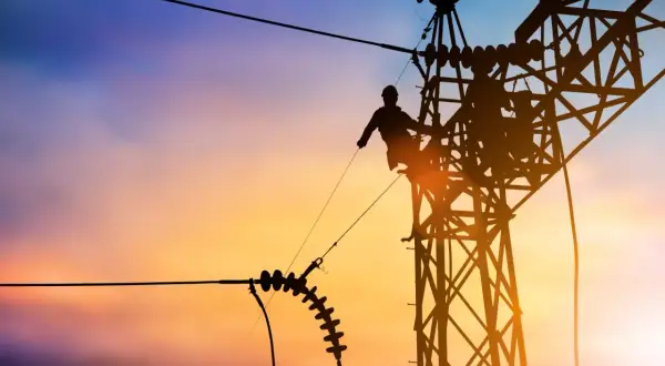 Sylwetka pracownika na tle zachodzącego słońca, naprawiającego linię energetyczną. Widać izolatory i konstrukcję wieży, a także potencjalny łuk elektryczny.