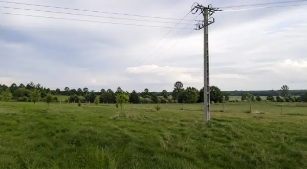 Słup na działce ponad 30 lat, stoi pośród zielonej łąki, z liniami energetycznymi biegnącymi ku horyzontowi.