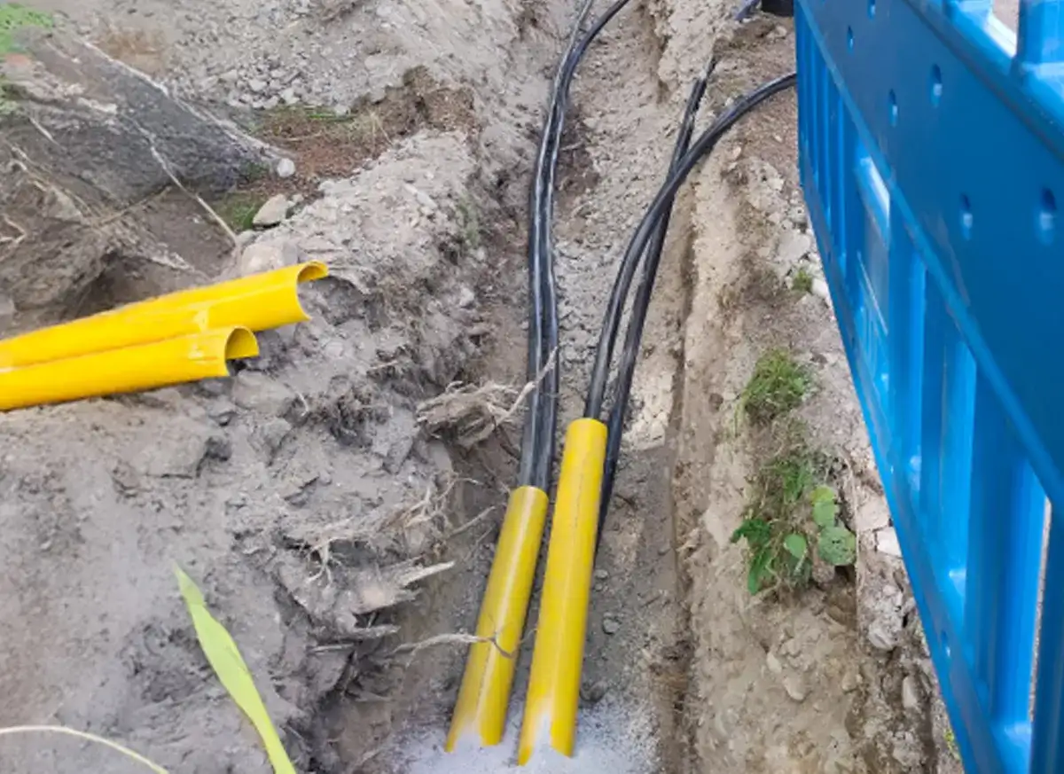 Wykop pod instalację światłowodową. Widoczne żółte osłony kabli i czarne przewody w wykopie. Obok niebieskie ogrodzenie budowlane.