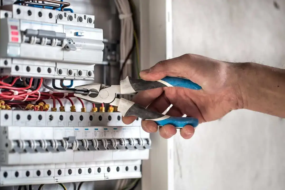 Ręka z kombinerkami przy skrzynce z bezpiecznikami. Gdy nastąpi brak prądu, gdzie dzwonić? Elektryk naprawia instalację.